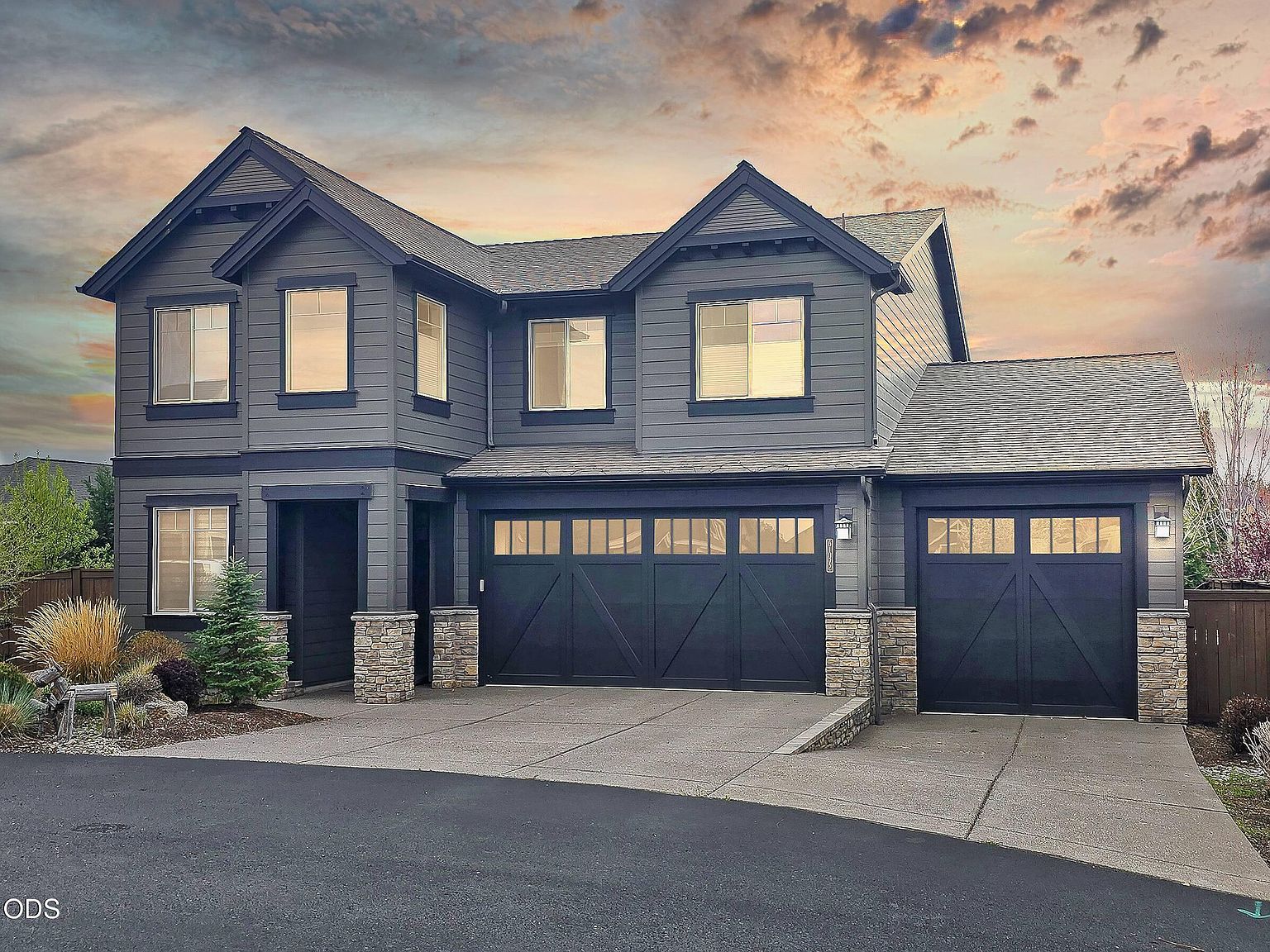 Twilight exterior of 61072 Manhae Loop, a 2015 Pahlisch Craftsman with stone-base columns, board-and-batten siding, and a four-car garage with carriage doors, set against a pink-orange Cascade sky.